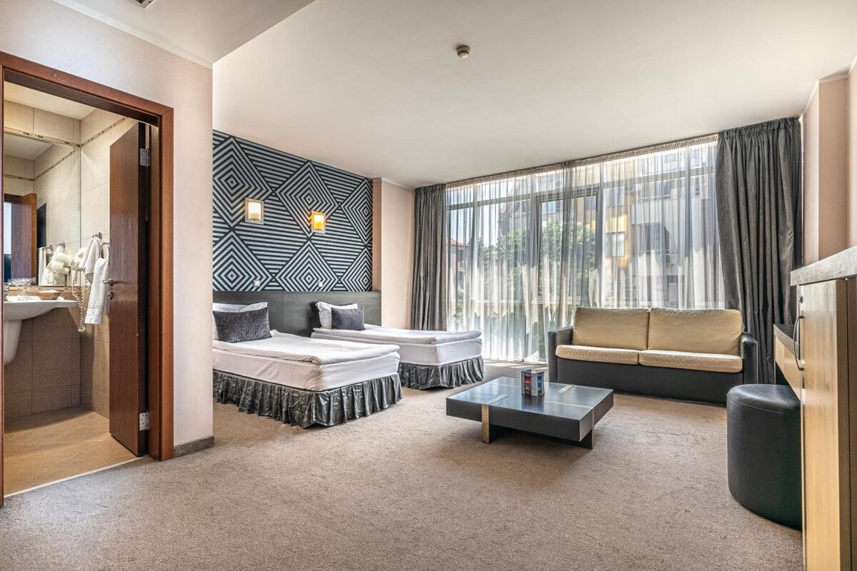 A hotel room with a bed, a sofa, a coffee table, and a large window with curtains.