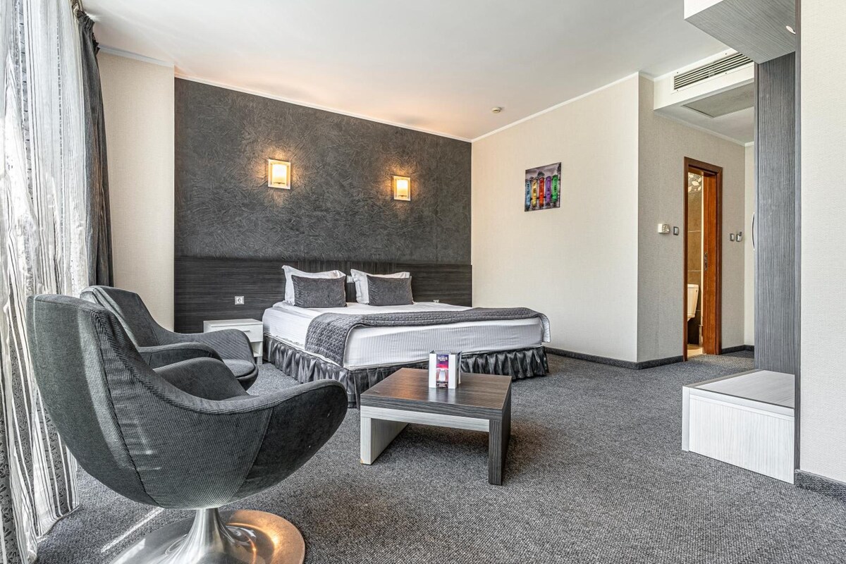 A hotel room with a bed, a chair, a small table, and a painting on the wall.