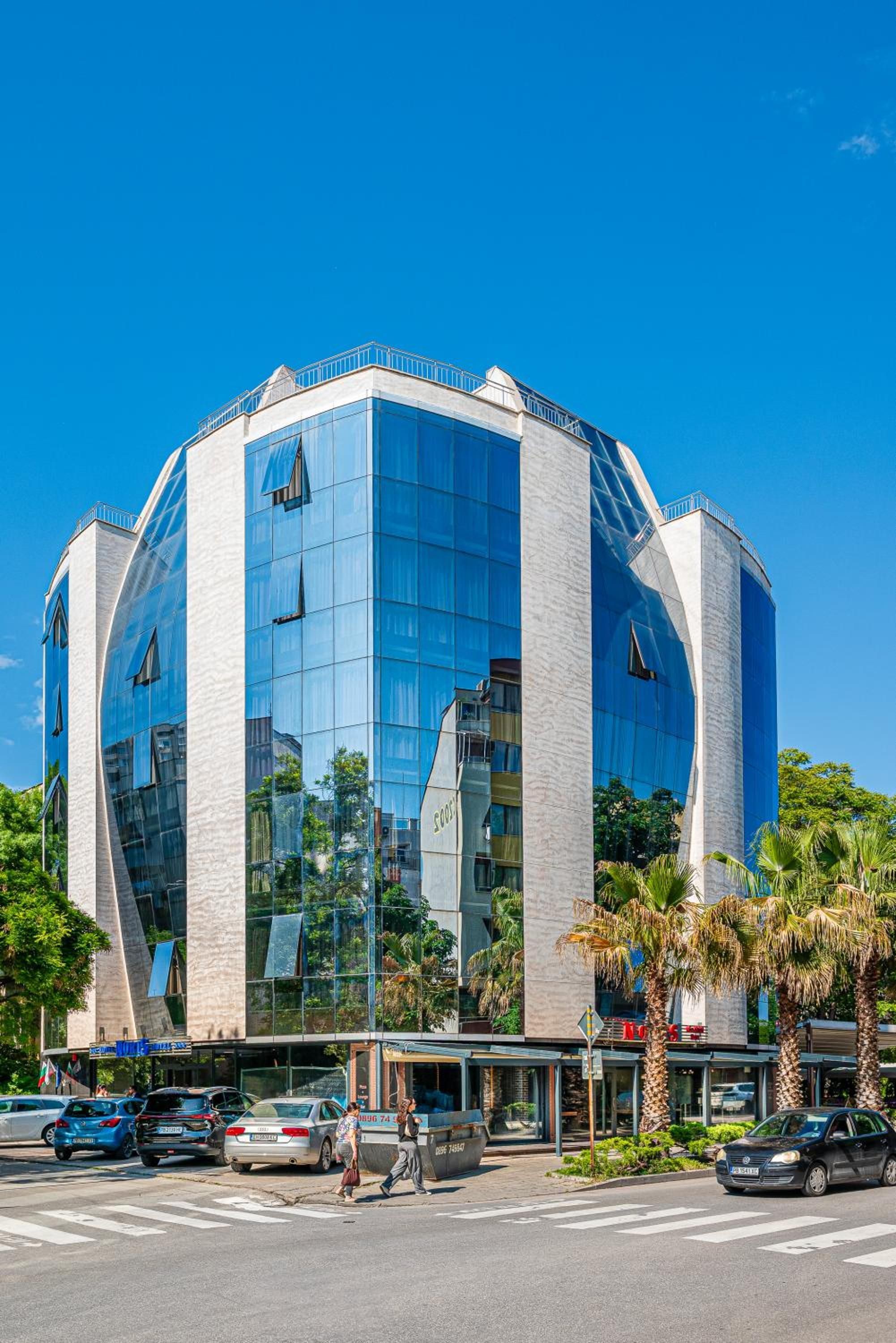 A modern hotel with a glass facade, a central courtyard, and a parking area with cars.
