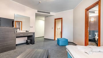 A hotel room with a bed, a desk with a mirror, a small blue ottoman, and a bathroom visible through an open door.