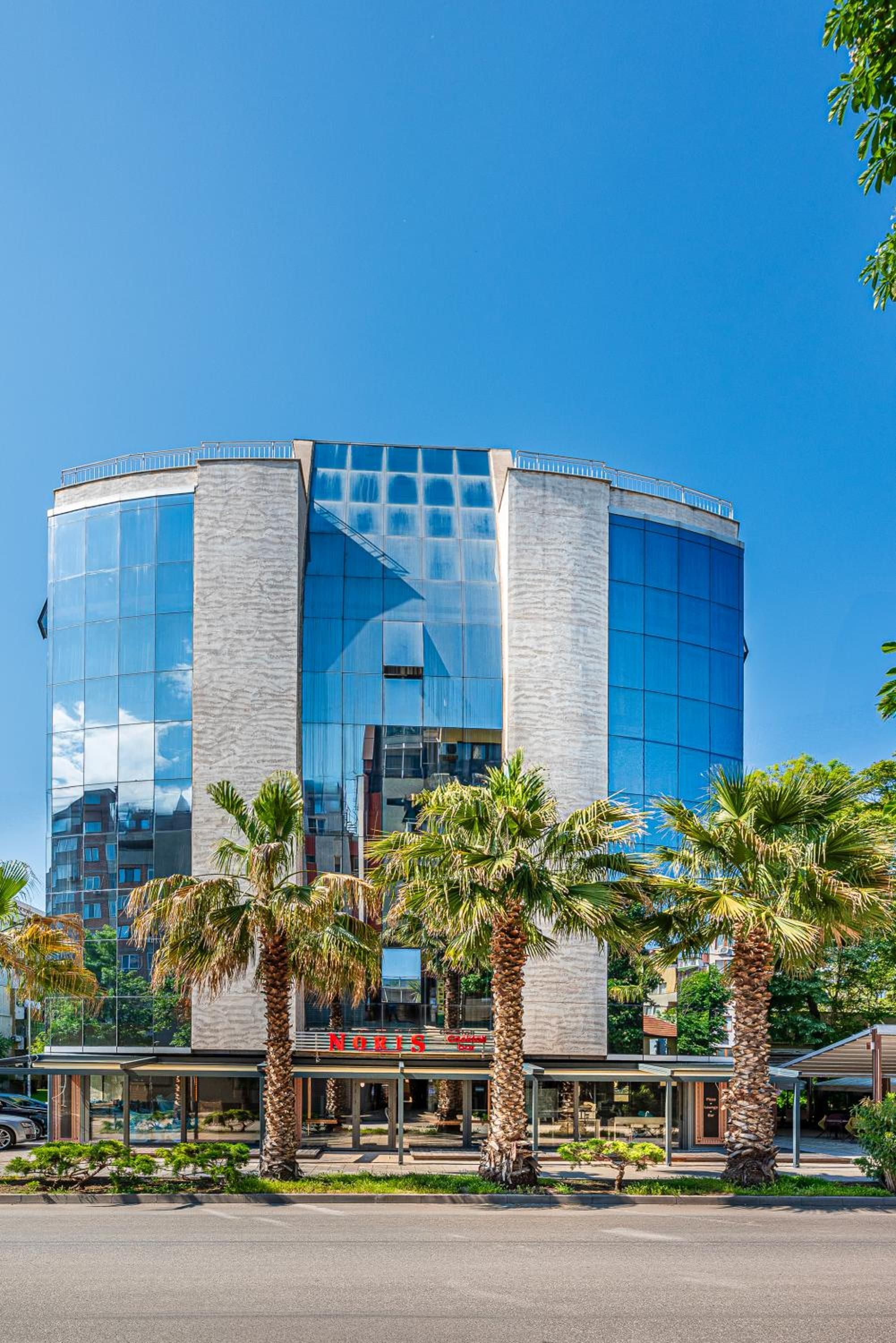 A modern building with a glass facade, palm trees in the foreground, and a clear blue sky.