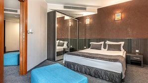 A hotel room with a large bed, a bedside table, a nightstand, a mirror, and a blue ottoman.