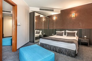 A hotel room with a large bed, a bedside table, a nightstand, a mirror, and a blue ottoman. - Hotel Noris (Plovdiv)