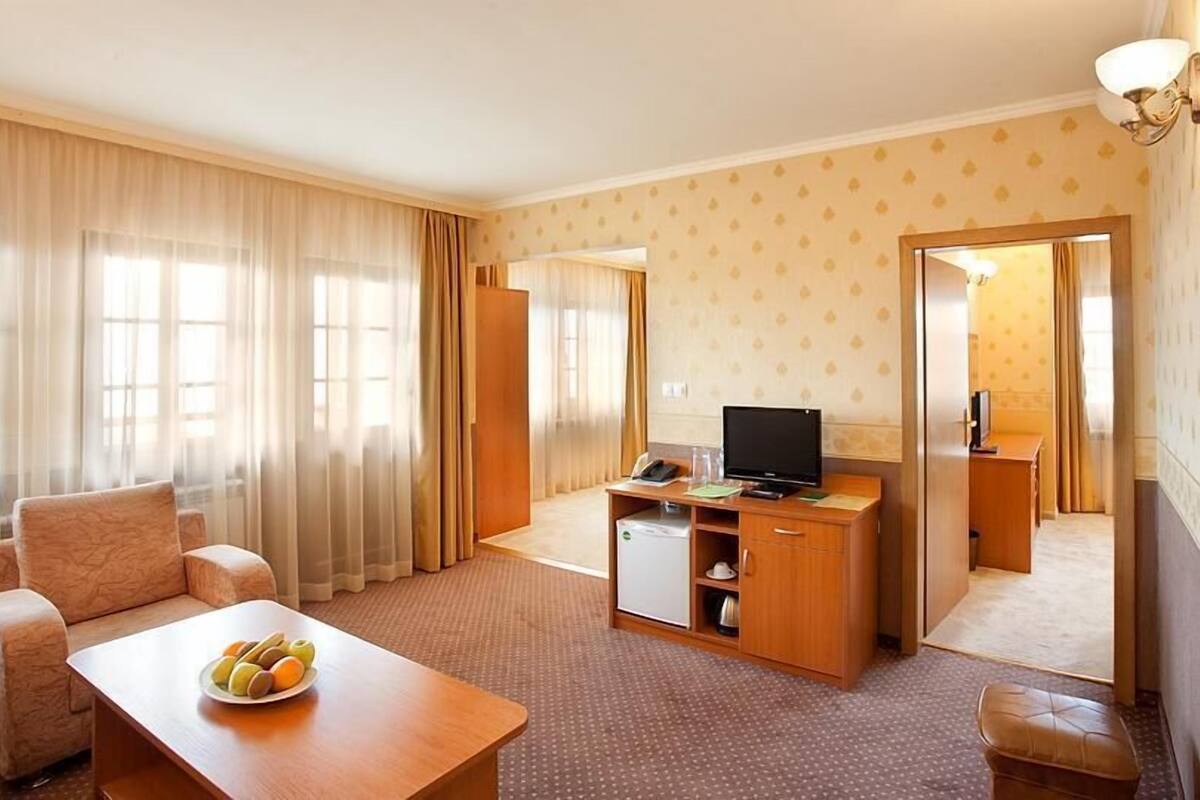 A hotel room with a sofa, a dining table with fruit, a TV, a mini-fridge, and a mirror.