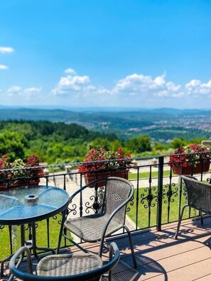 Une terrasse avec une table ronde en verre et deux chaises en osier, offrant une vue panoramique sur un paysage vallonné sous un ciel dégagé.