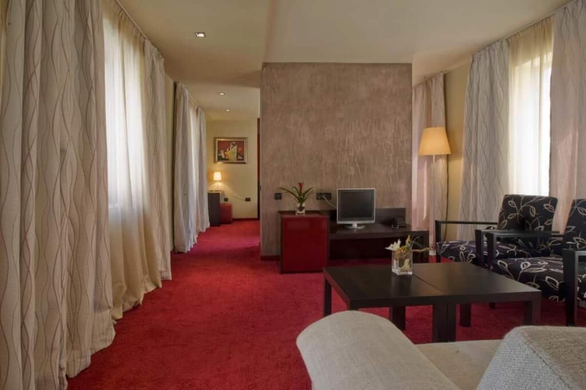 Une chambre d’hôtel avec un tapis rouge, une télévision, une petite table avec un vase, un canapé et un fauteuil.