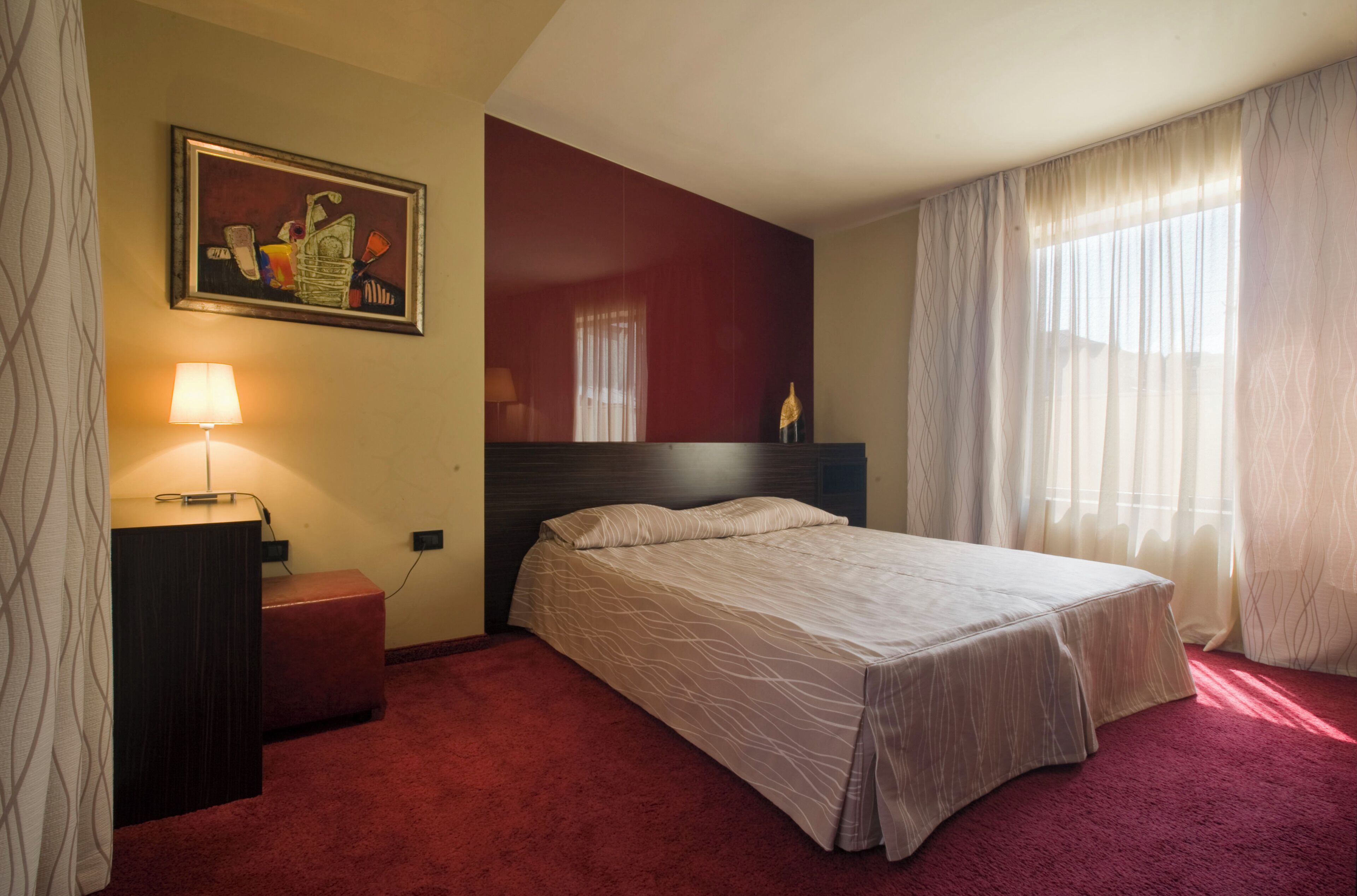 A hotel room with a bed, a nightstand, a lamp, a painting, and a window with curtains.