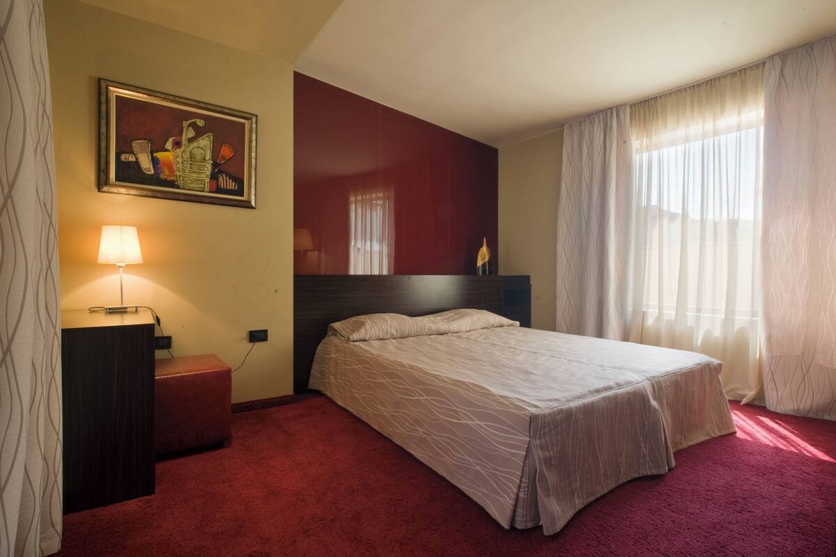 Une chambre d’hôtel avec un lit, une table de chevet, une lampe, un tableau et une fenêtre avec des rideaux.