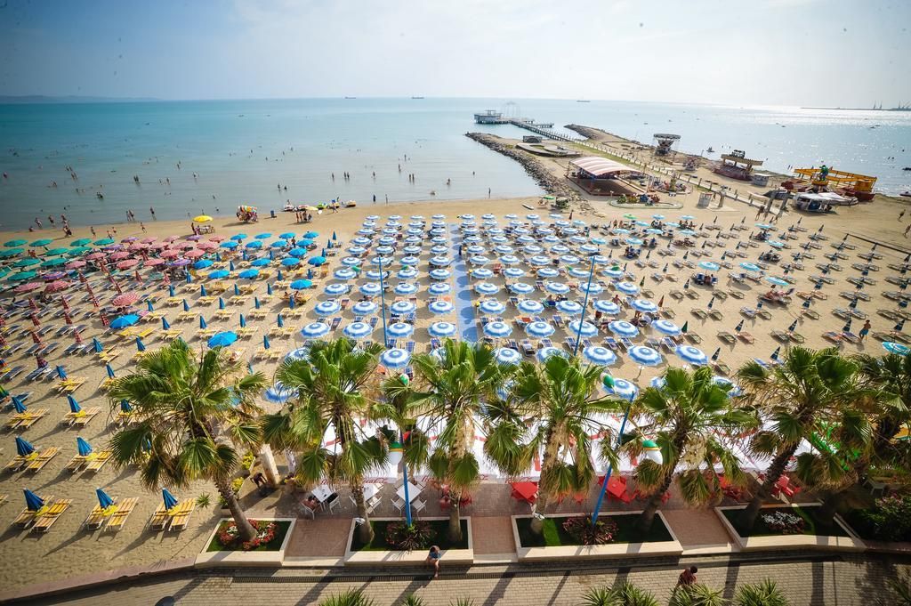 Ein Strand mit vielen Sonnenschirmen und Liegestühlen, Palmen und einem Pier im Hintergrund.