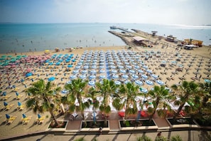A beach with numerous umbrellas and sun loungers, palm trees, and a pier in the background. - Iliria International (Durrës)