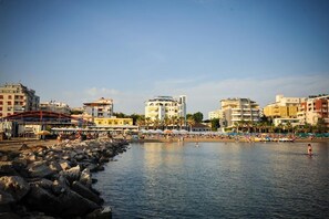 A coastal cityscape with buildings, a rocky shoreline, and a beach with people. - Iliria International (Durrës)