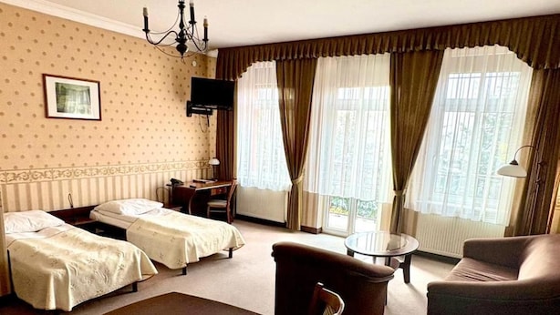 Ein Hotelzimmer mit Esstisch, zwei Einzelbetten, einer Couch, einem Fernseher und einem Fenster mit Vorhängen.