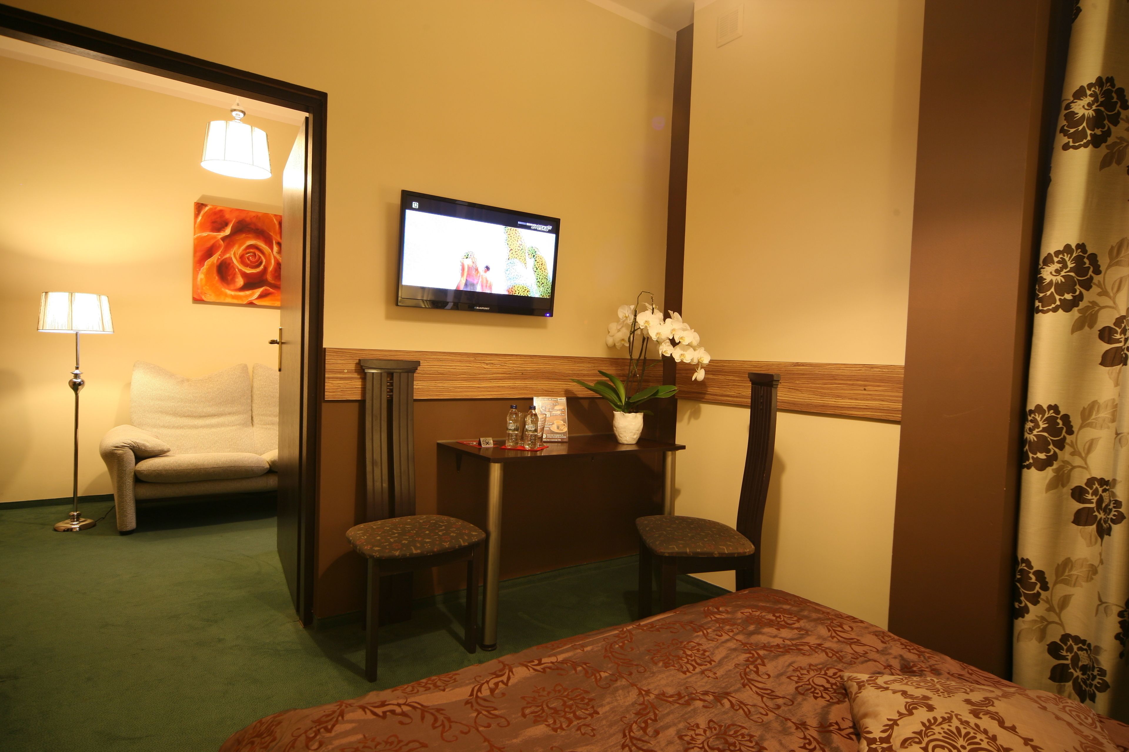 A hotel room with a bed, a desk, two chairs, a TV, a potted plant, and a painting on the wall.
