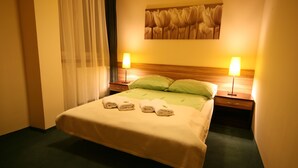 A hotel room with a bed, two bedside lamps, a green pillow, a headboard with a floral painting, and a window with sheer curtains. - Hotel Seven 7 (Kalisz)