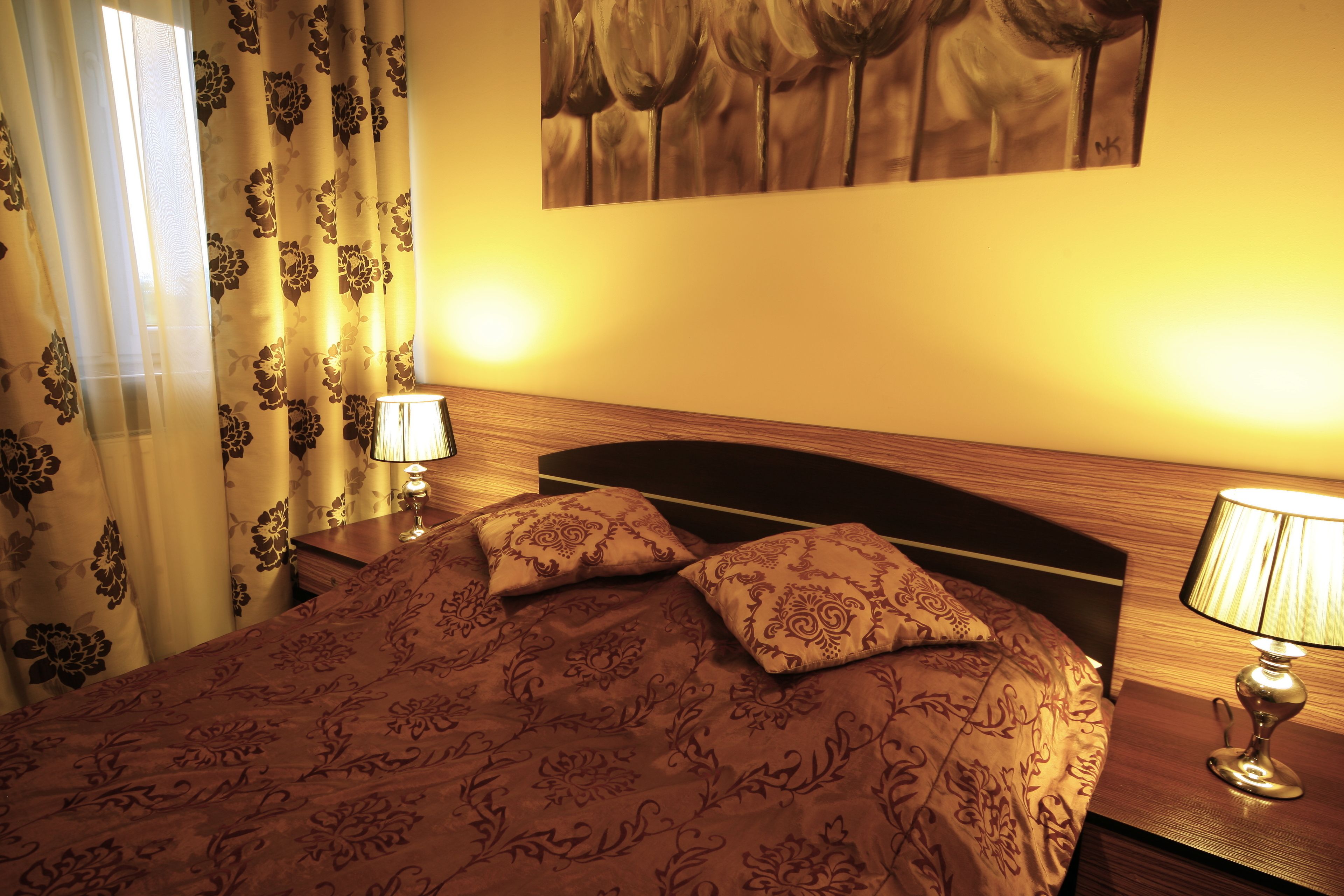 Un lit bien fait, avec une couvre-lit à motifs et deux oreillers, une table de chevet avec une lampe, et un tableau accroché au mur.