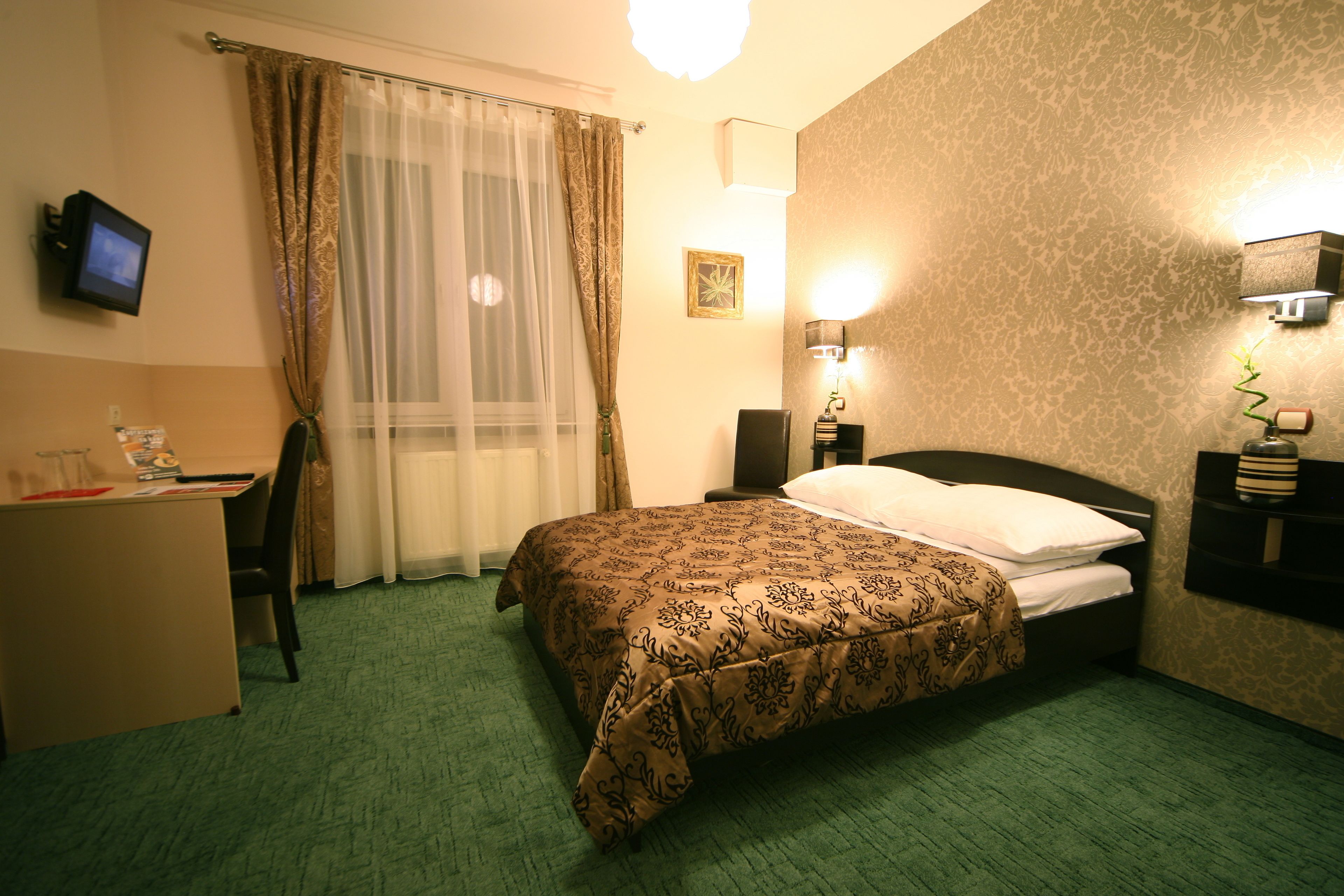 Quarto de hotel com cama, escrivaninha, cadeira, televisão e luminária na parede.