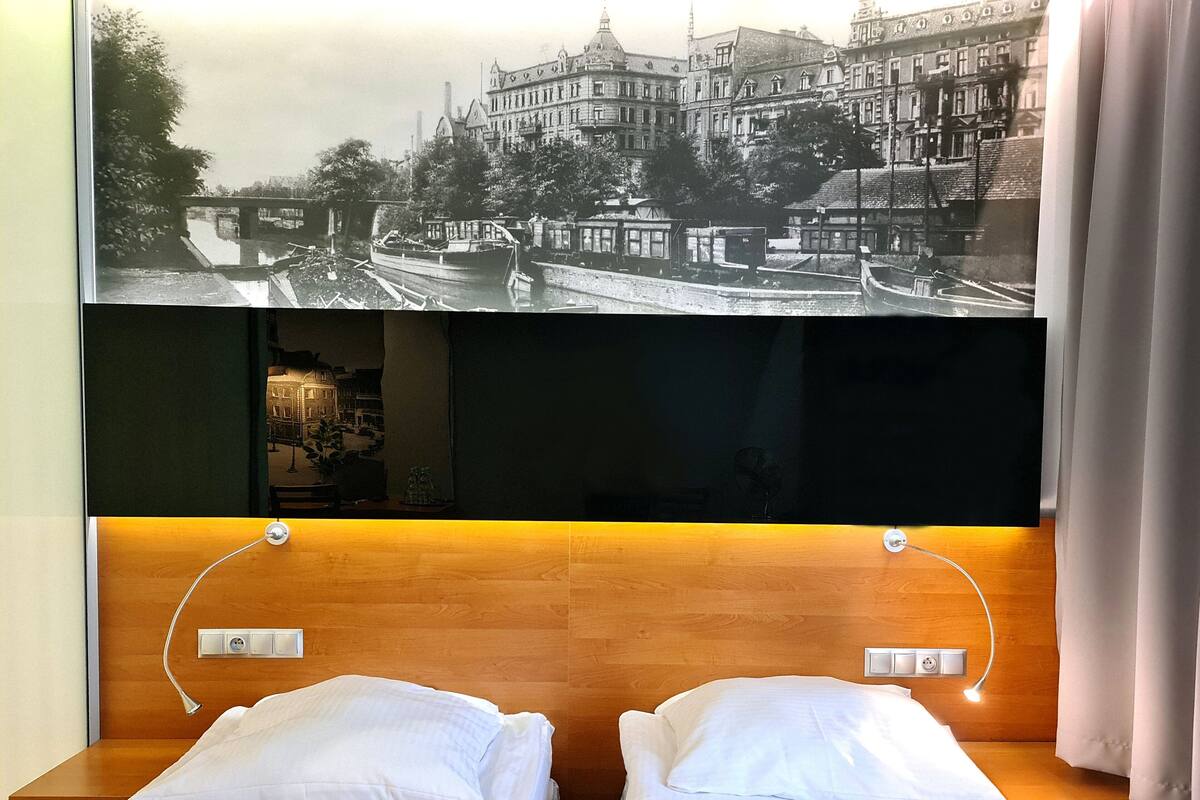A hotel room with two single beds, a large flat-screen TV mounted above the beds displaying a black and white photo, and a wooden headboard.