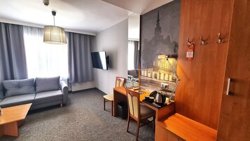 Una habitación de hotel con sofá, una mesa de comedor con sillas, un televisor montado en la pared y un cuadro de un paisaje urbano.