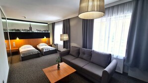 Quarto de hotel com sofá, duas camas de solteiro, uma mesa de centro de madeira e um mural com vista urbana na parede.