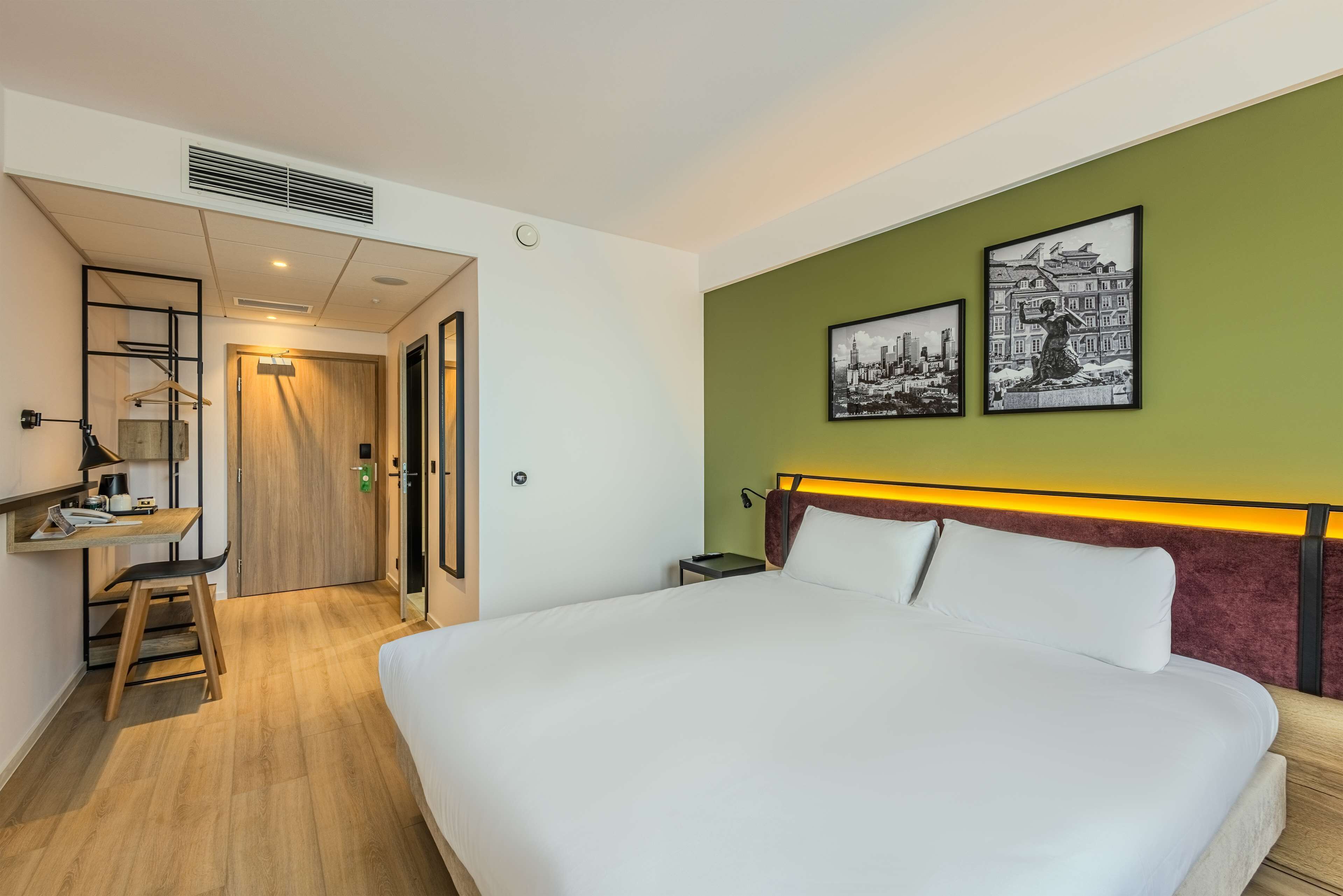 A hotel room with a large bed, a desk with a chair, a wardrobe, and two framed pictures on the wall.
