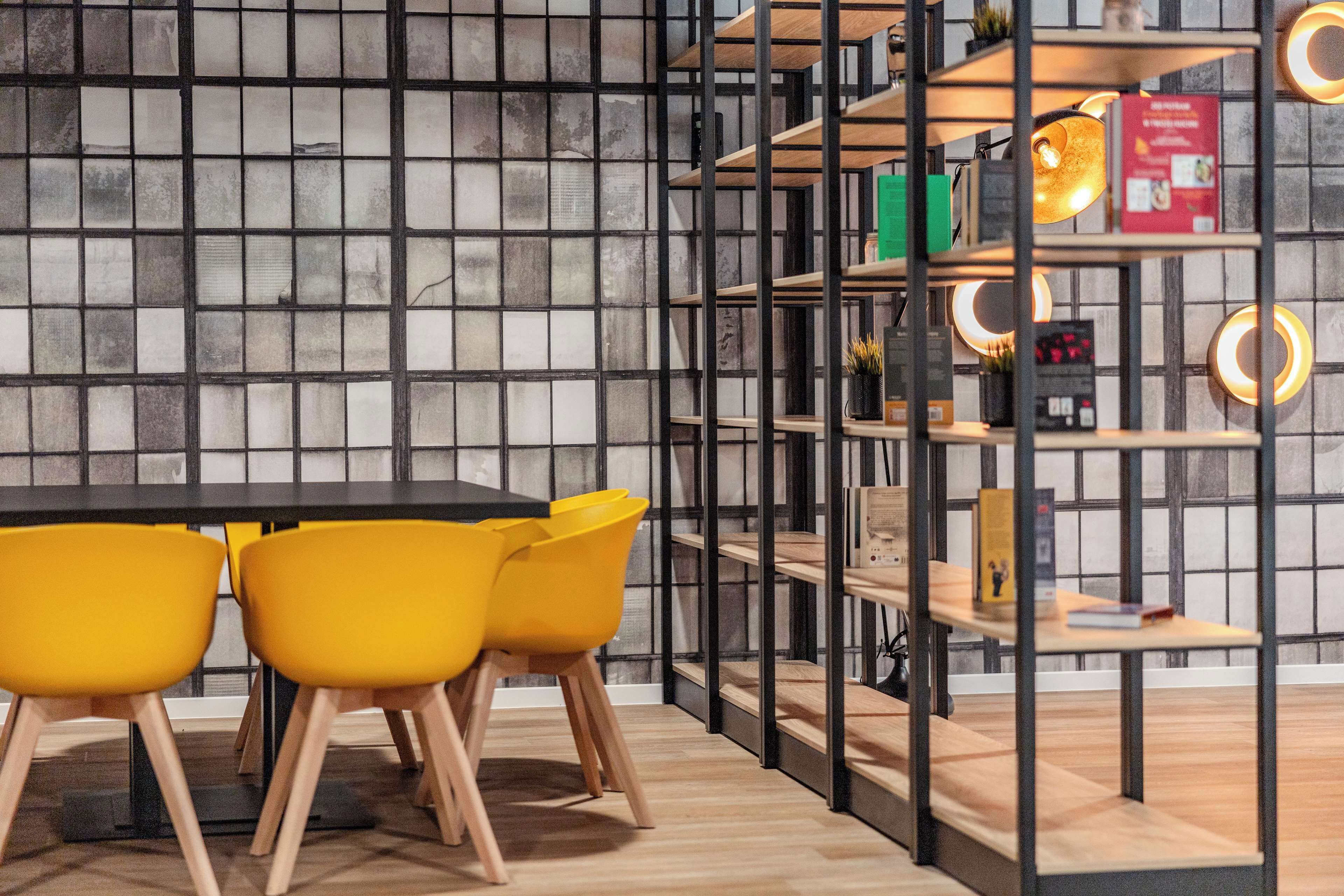 A modern interior with a black and white tiled wall, a wooden table with yellow chairs, and a metal shelving unit with books and decorative items.