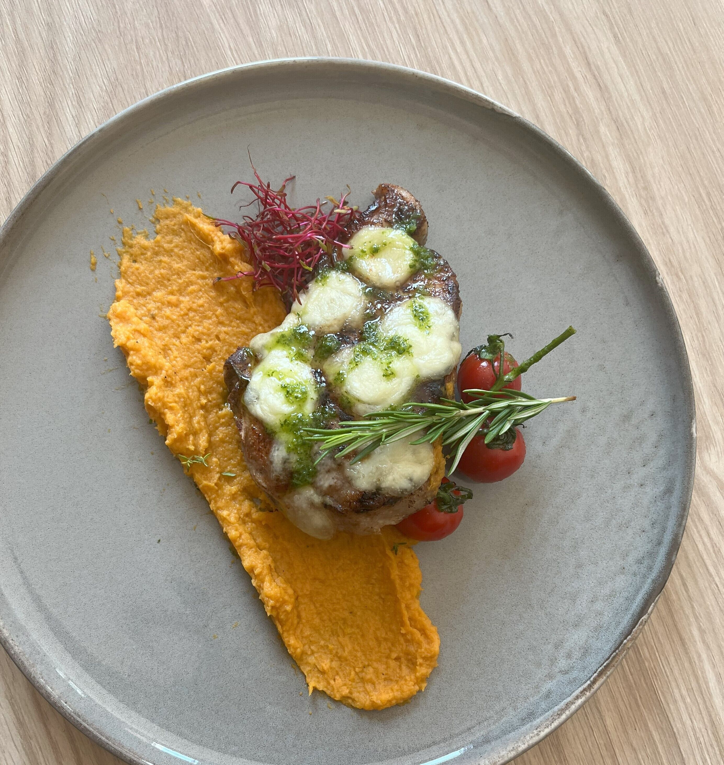 Een bord met een steak, belegd met kaas en kruiden, vergezeld van een portie aardappelpuree en kersen-tomaten.