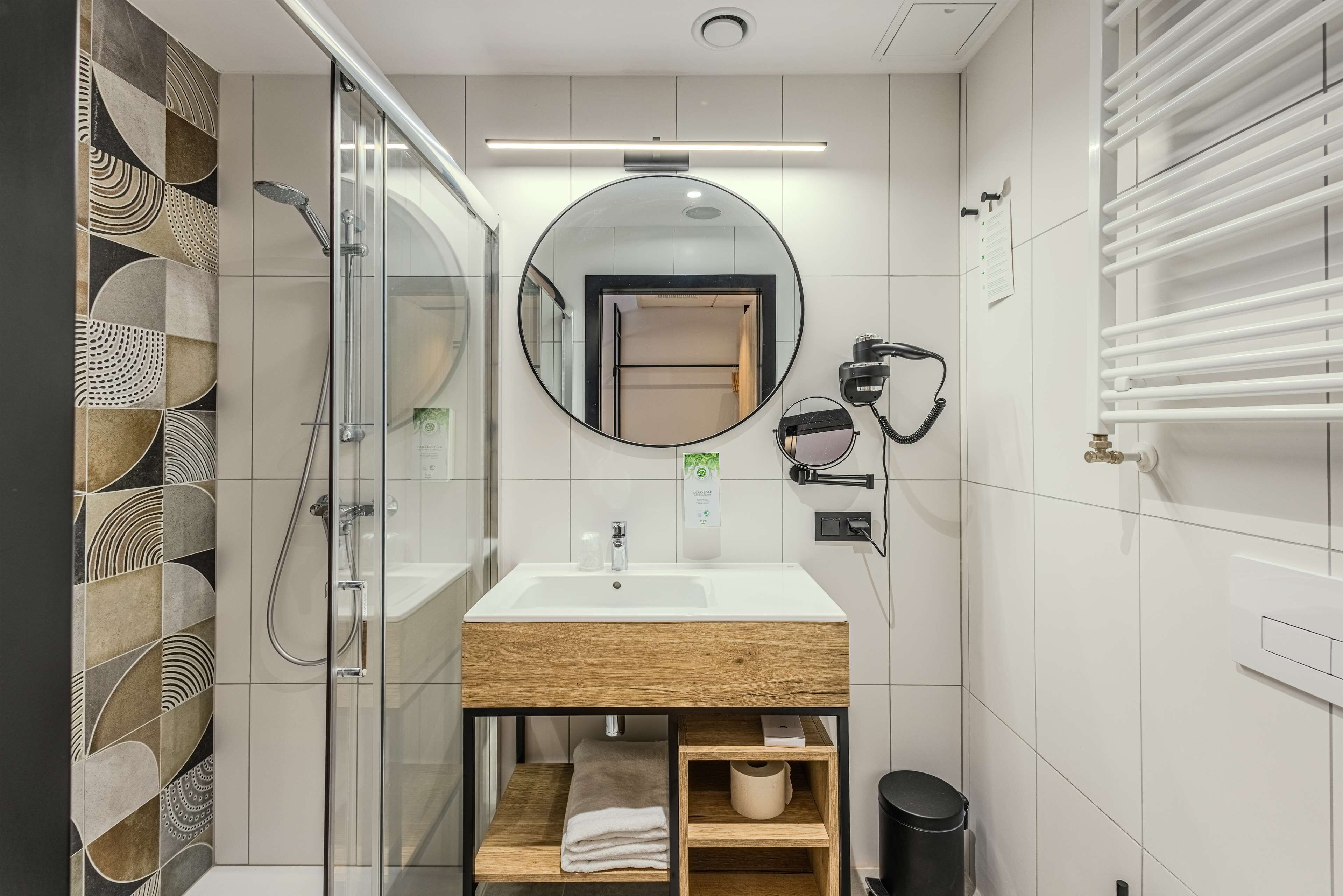 A modern bathroom with a glass shower enclosure, a wooden vanity, a round mirror, and a towel rack.