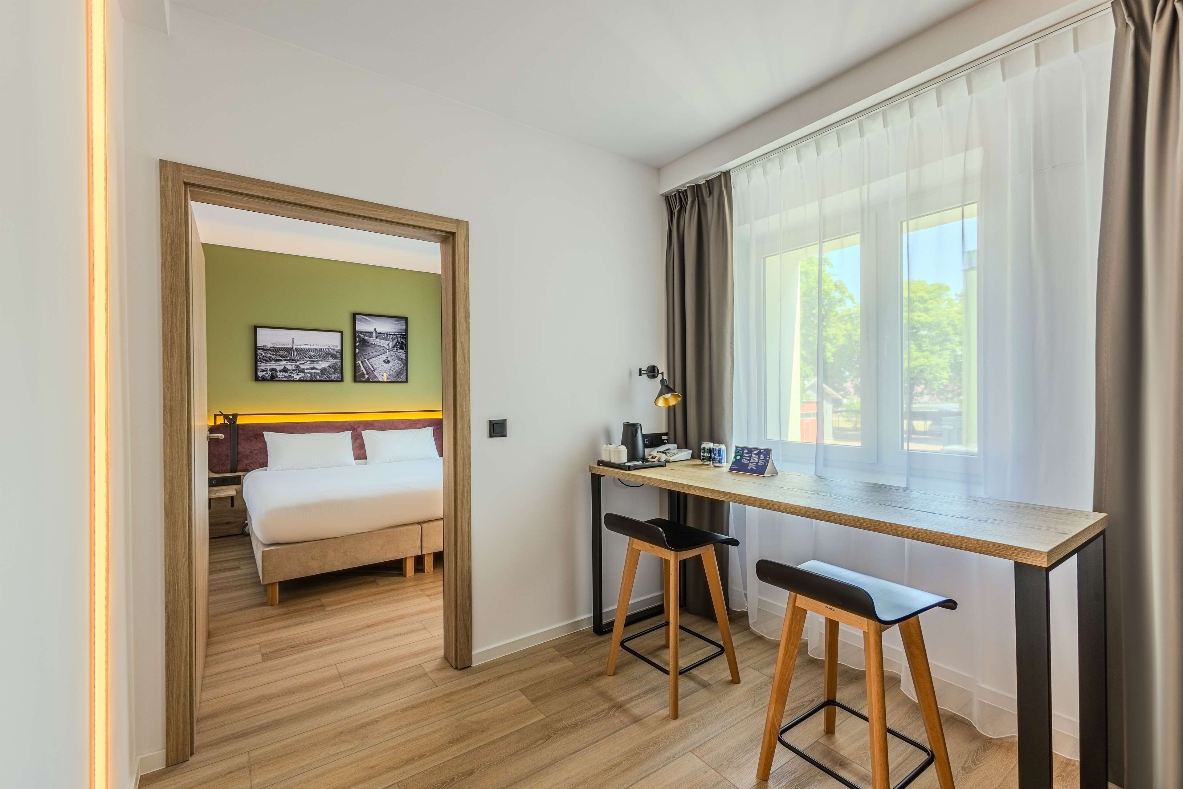Habitación de hotel con cama, escritorio con dos sillas, ventana con cortinas y dos cuadros en la pared.