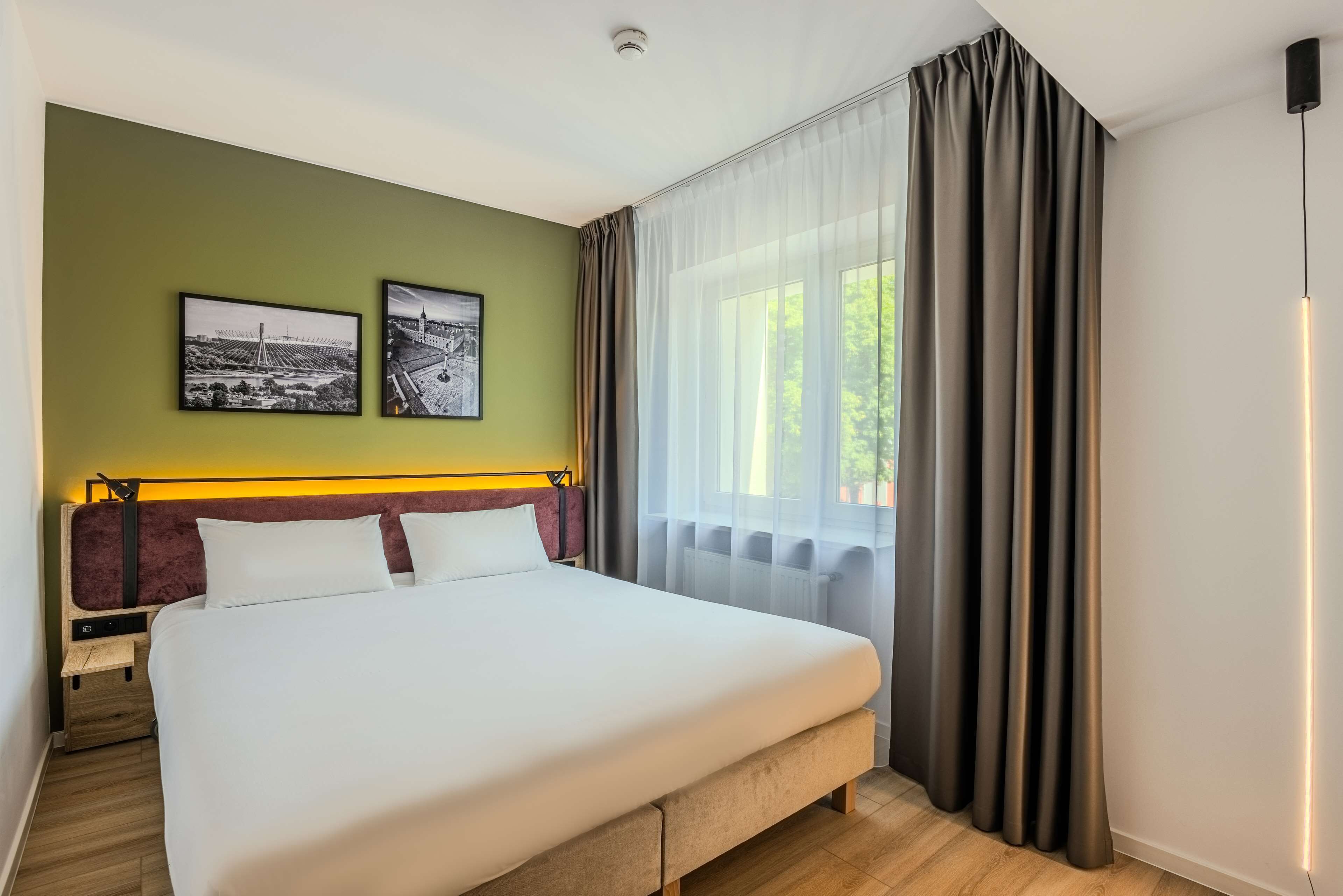 Ein Hotelzimmer mit einem großen Bett, zwei gerahmten Bildern an der Wand, einem Fenster mit Vorhängen und einem Kopfteil mit einer Leuchte.
