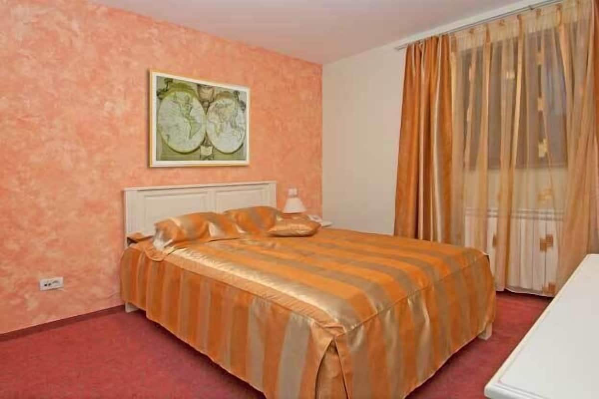 Hotellihuone, jossa on sänky, yöpöytä, lamppu, ikkuna verhoilla ja seinä, jossa on kehystetty taulu.