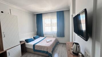 Un dormitorio con cama, televisor, escritorio con hervidor y ventana con cortinas.