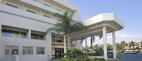 A modern hotel with a covered entrance, multiple floors, and a landscaped garden with palm trees.