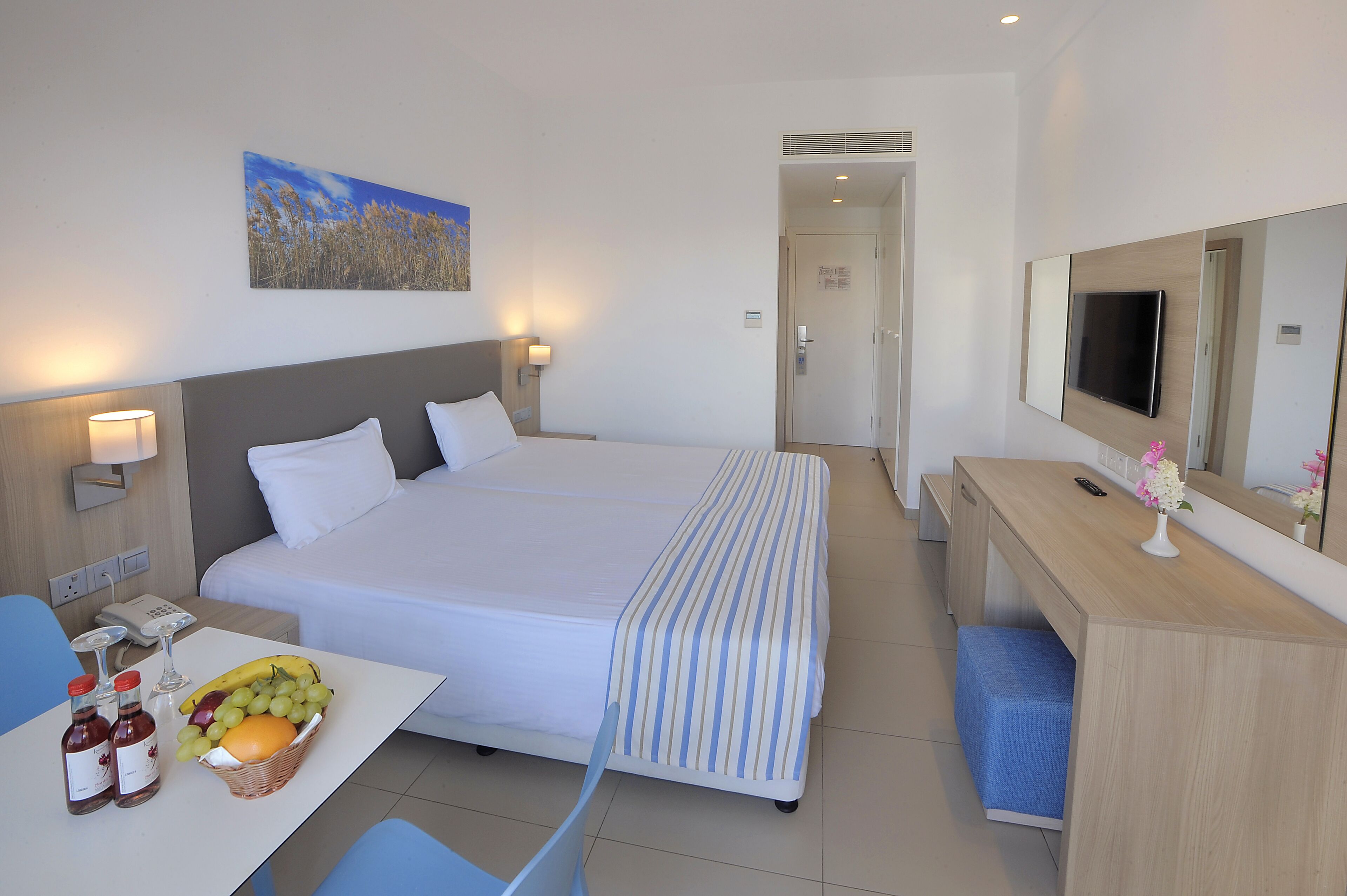 A hotel room with a large bed, bedside tables with lamps, a desk with a mirror, a TV, and a fruit basket.