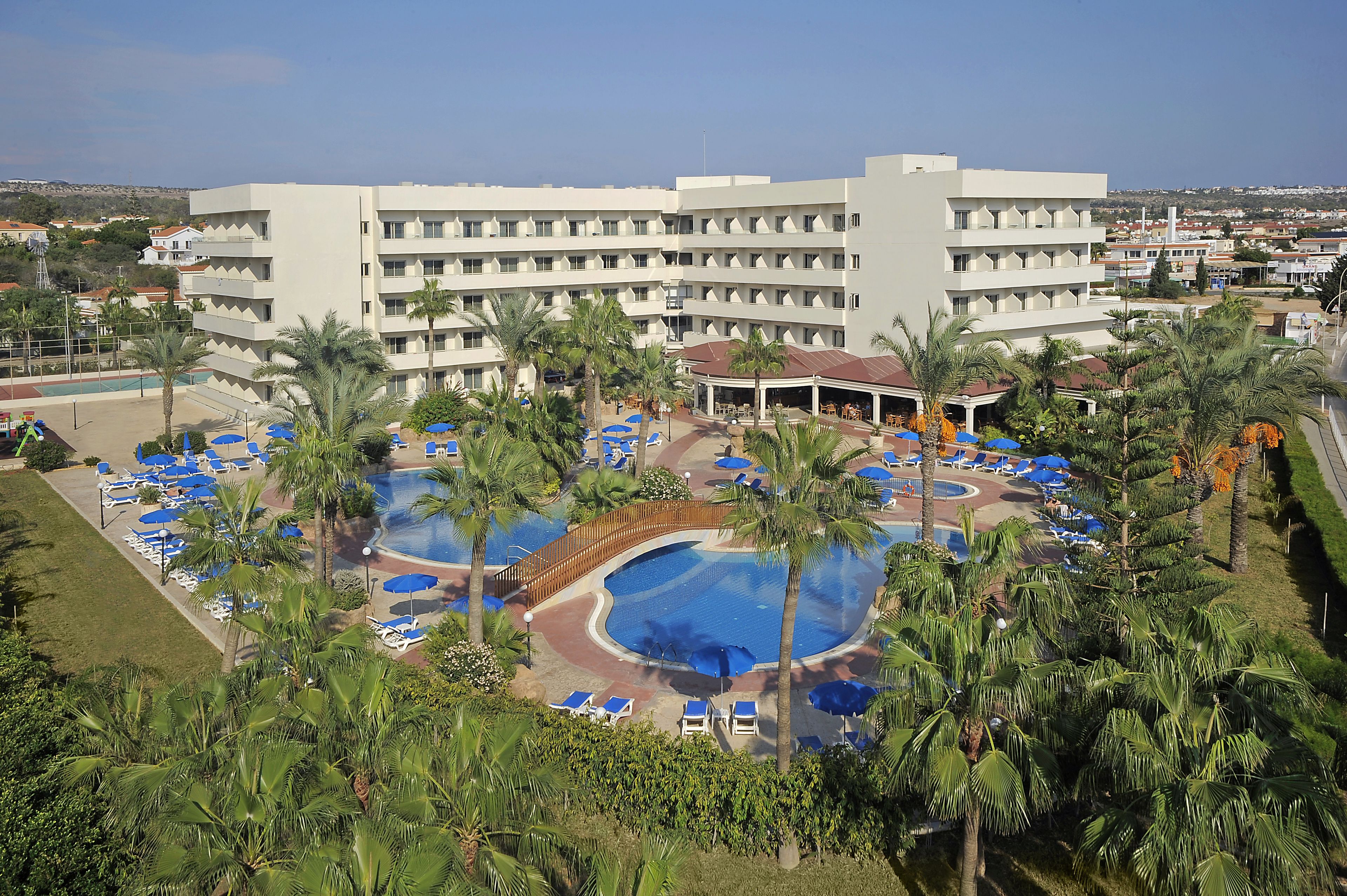 A hotel with a large pool area surrounded by palm trees and blue umbrellas, a covered seating area, and a view of the surrounding town.