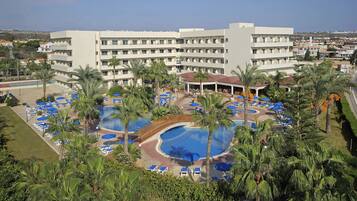 A hotel with a large pool area surrounded by palm trees and blue umbrellas, a covered seating area, and a view of the surrounding town.