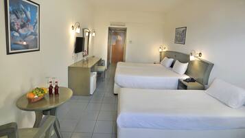 A hotel room with two beds, a desk with a computer, a small table with fruit and drinks, and artwork on the walls.