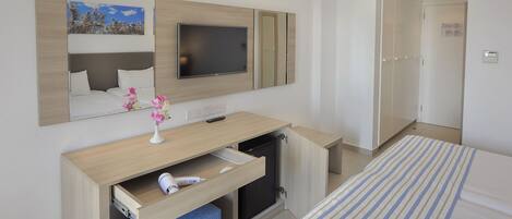 A hotel room with a bed, a desk with a TV, a mirror, and a small refrigerator.
