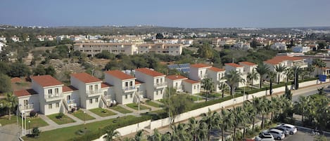 A residential area with multiple two-story buildings, each with a red-tiled roof and balconies. There is a parking lot with several cars and a few motorcycles. Palm trees line the streets, and the area appears to be well-maintained with green lawns and pathways.