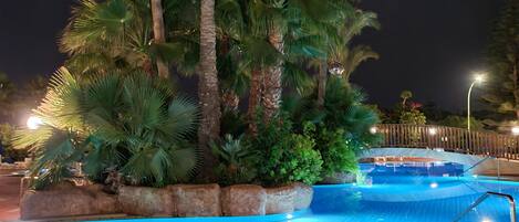 A nighttime swimming pool with palm trees and a wooden deck.