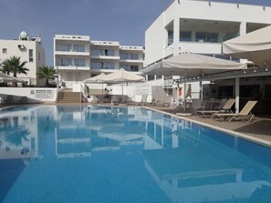 A swimming pool with lounge chairs and umbrellas, surrounded by modern buildings with balconies. - La Casa Di Napa (Ayia Napa)