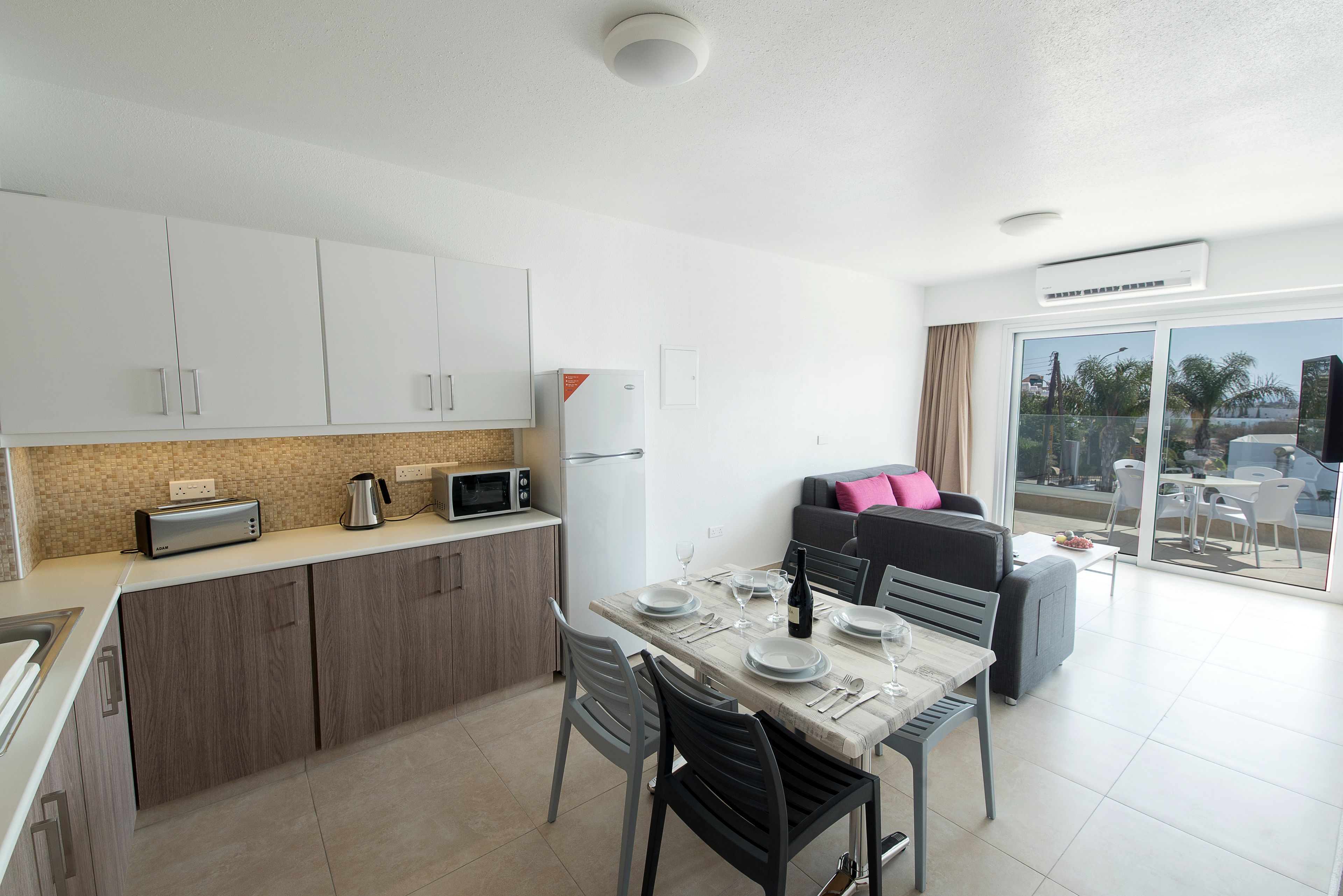 A modern kitchen with white cabinets, a dining table set for four, and a view of the outdoors through a sliding glass door.