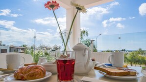 Una mesa con un croissant, un vaso de bebida roja, una tetera blanca y una taza, frente a una vista del mar y edificios.