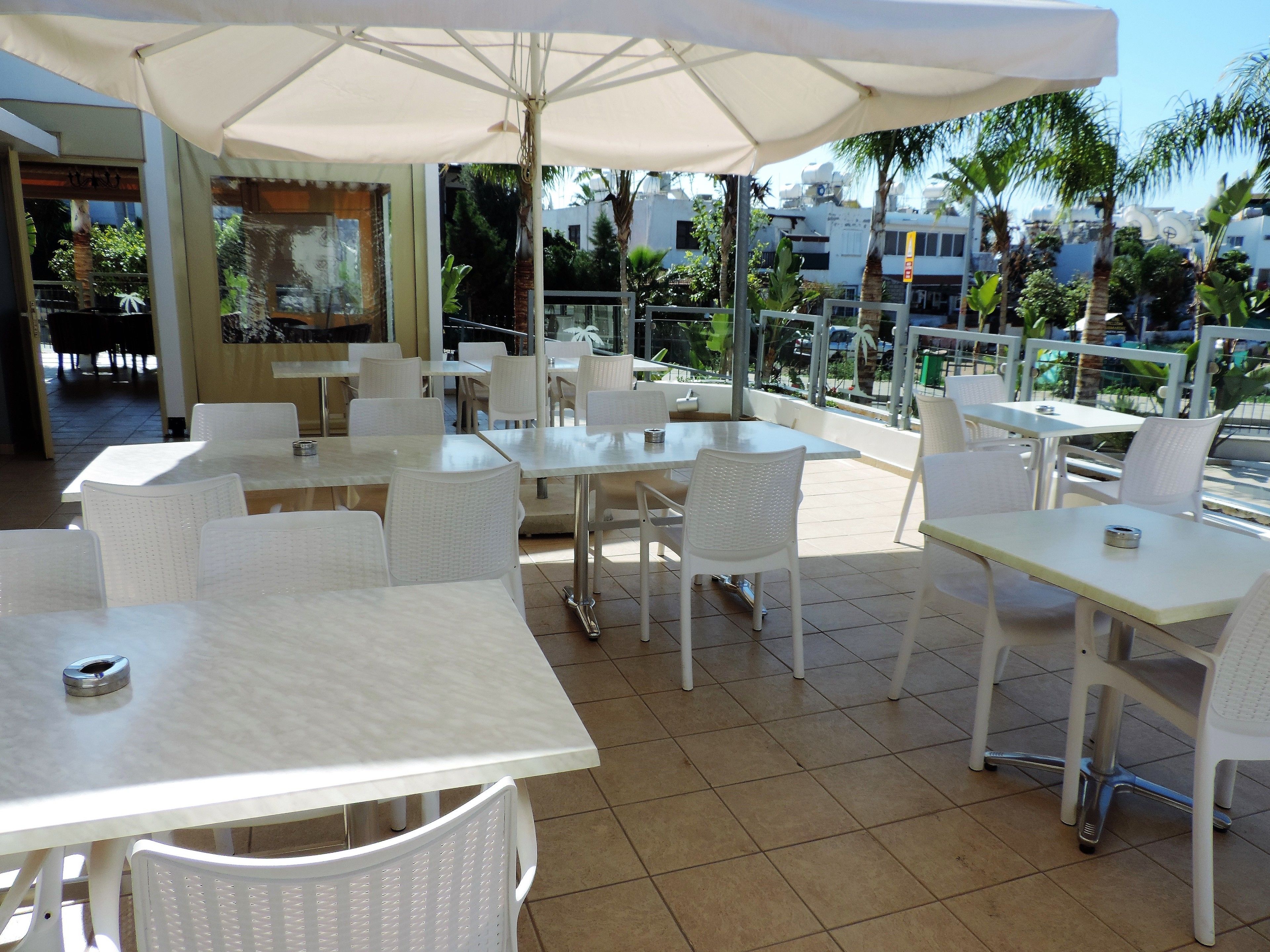 Un patio al aire libre con mesas y sillas blancas, sombrillas y vistas a palmeras y edificios.