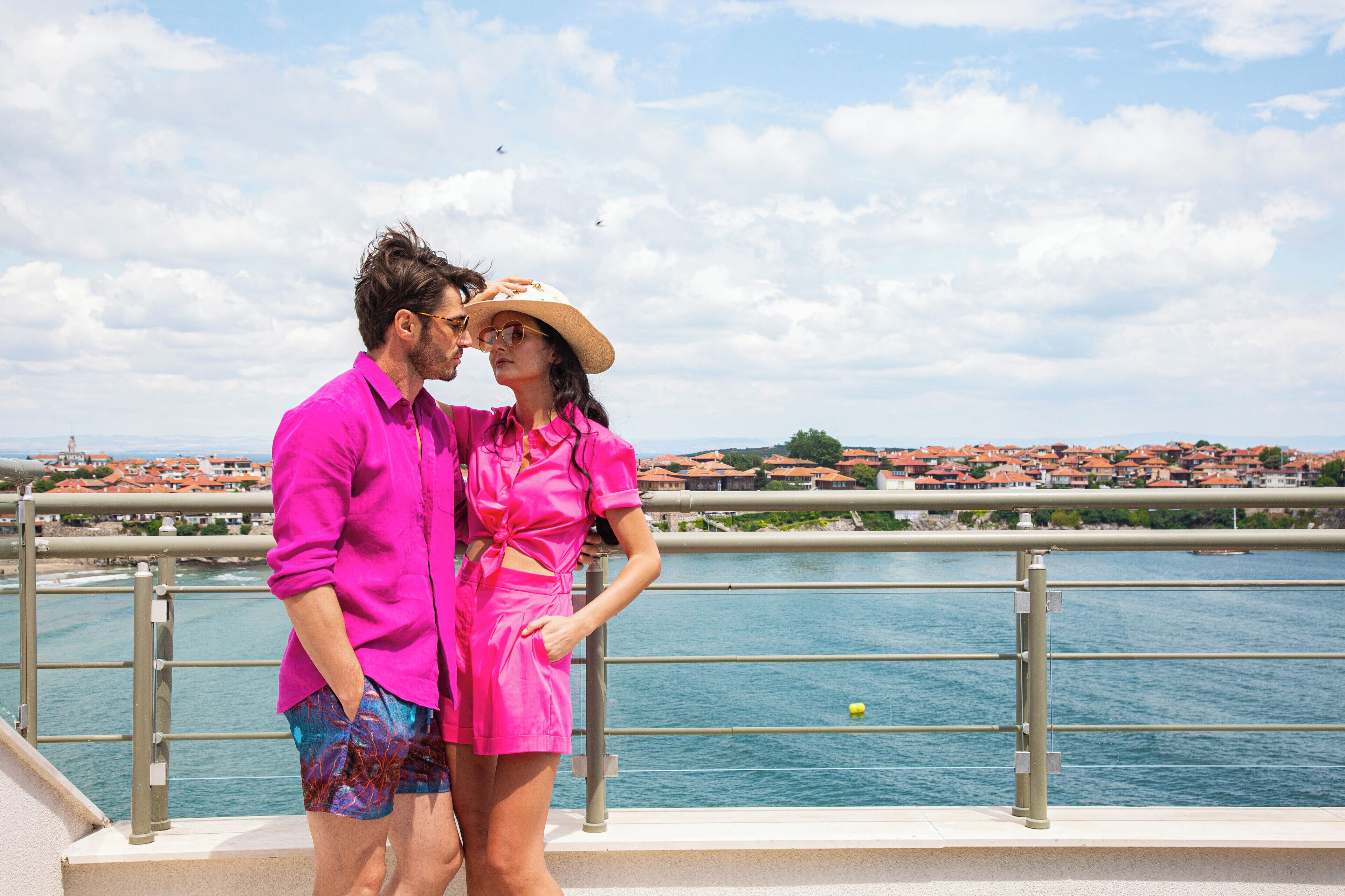 A couple standing on a balcony overlooking a coastal town with red-roofed buildings and a clear sky.