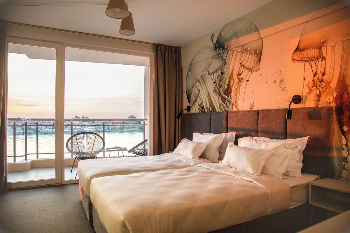 A hotel room with a large bed, a chair, a small table, and a view of the sea through a large window.