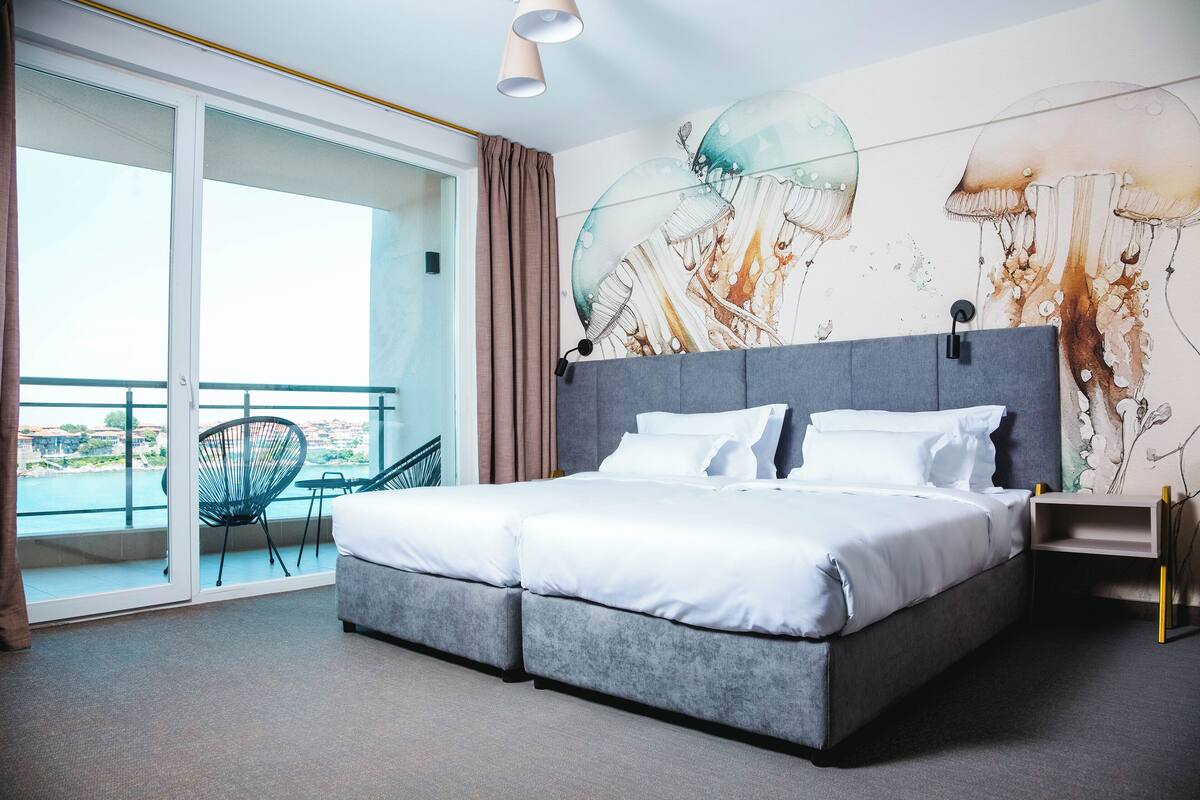 A modern hotel room with a large bed, a balcony with a chair, and a wall mural.