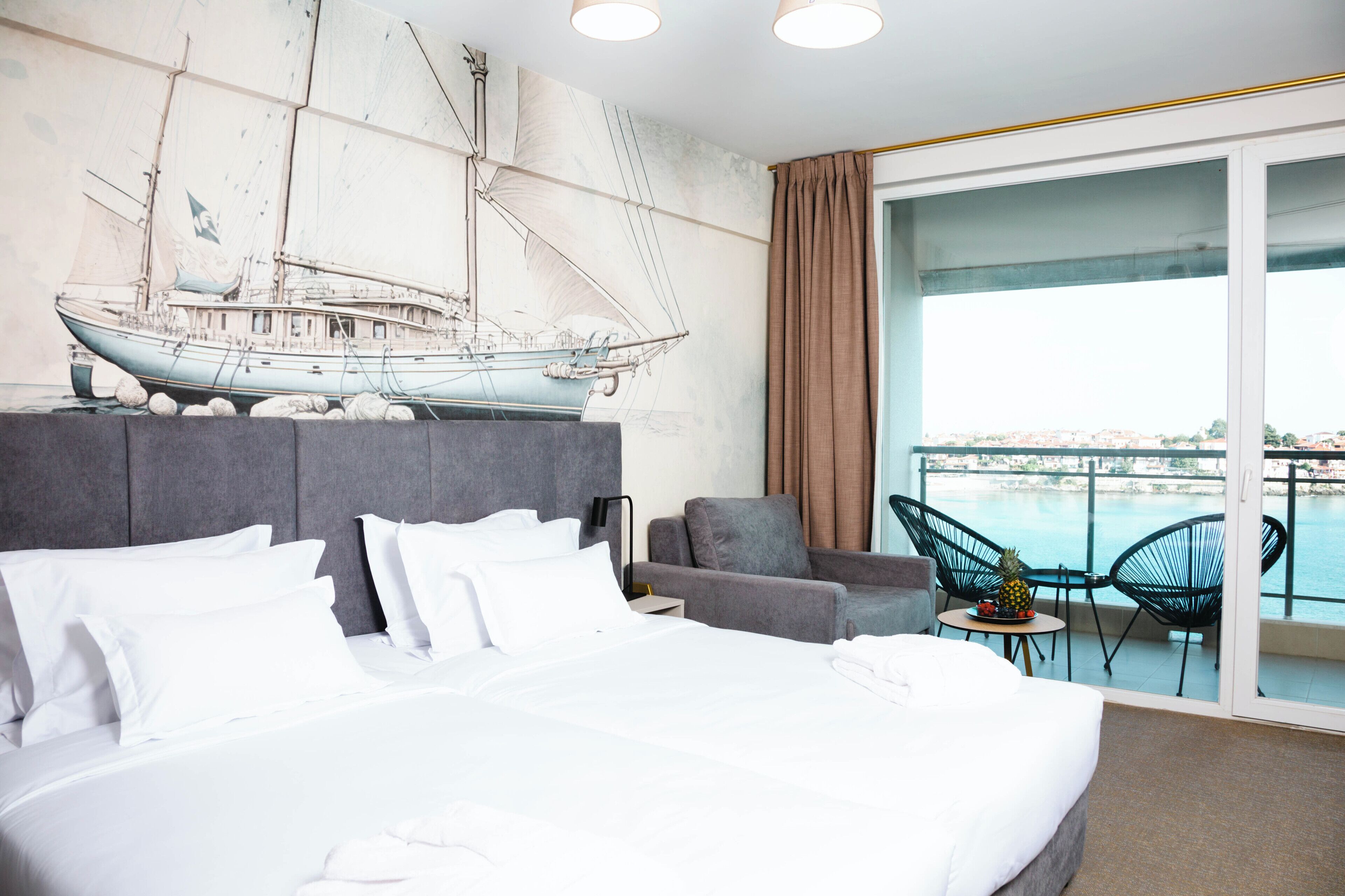 A hotel room with a large bed, a grey headboard, a wall-mounted ship illustration, a balcony with a view of the sea, and a small table with chairs.