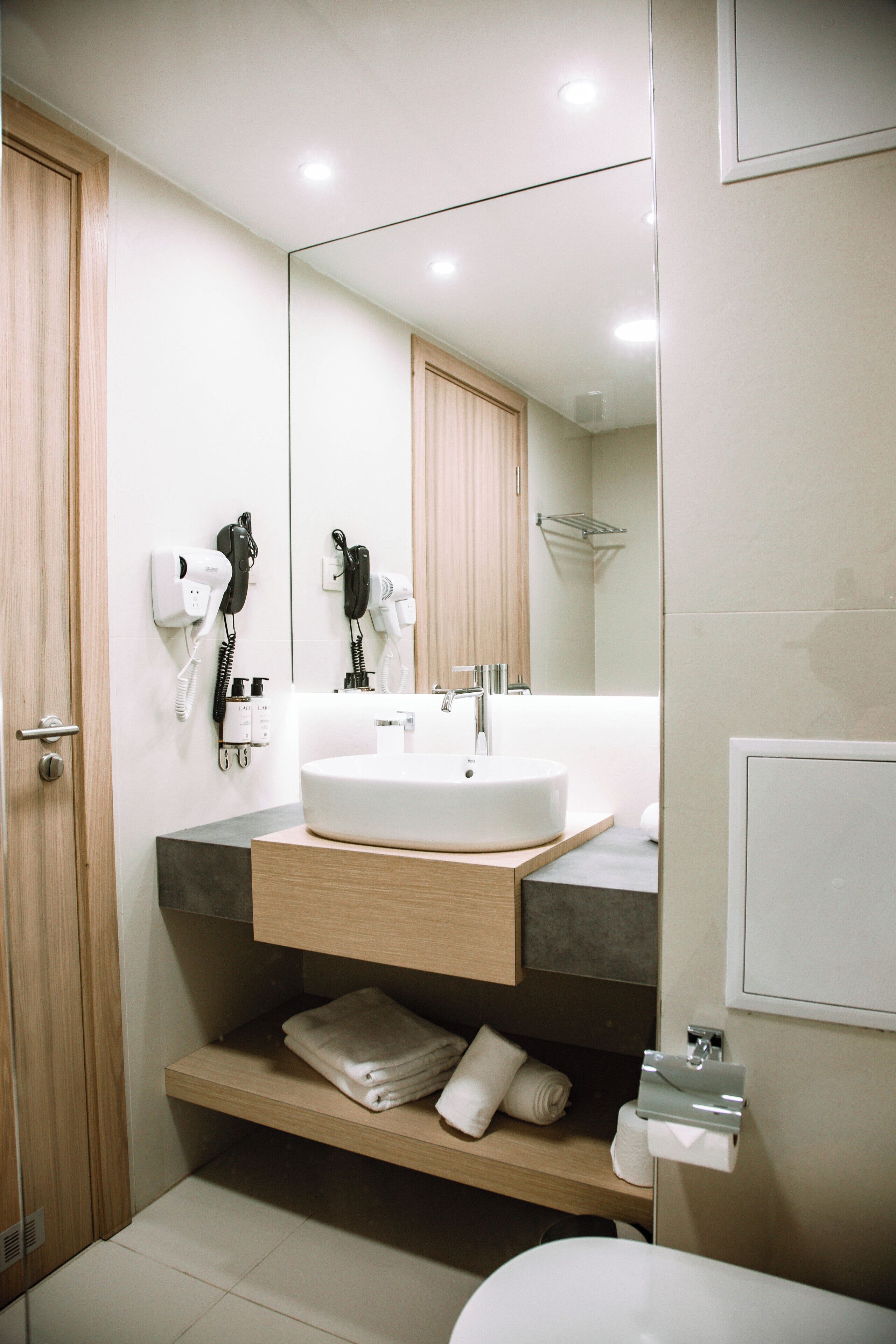 Une salle de bain moderne avec un lavabo blanc, un meuble-lavabo en bois et un miroir. On peut voir des sèche-cheveux et un porte-serviettes.