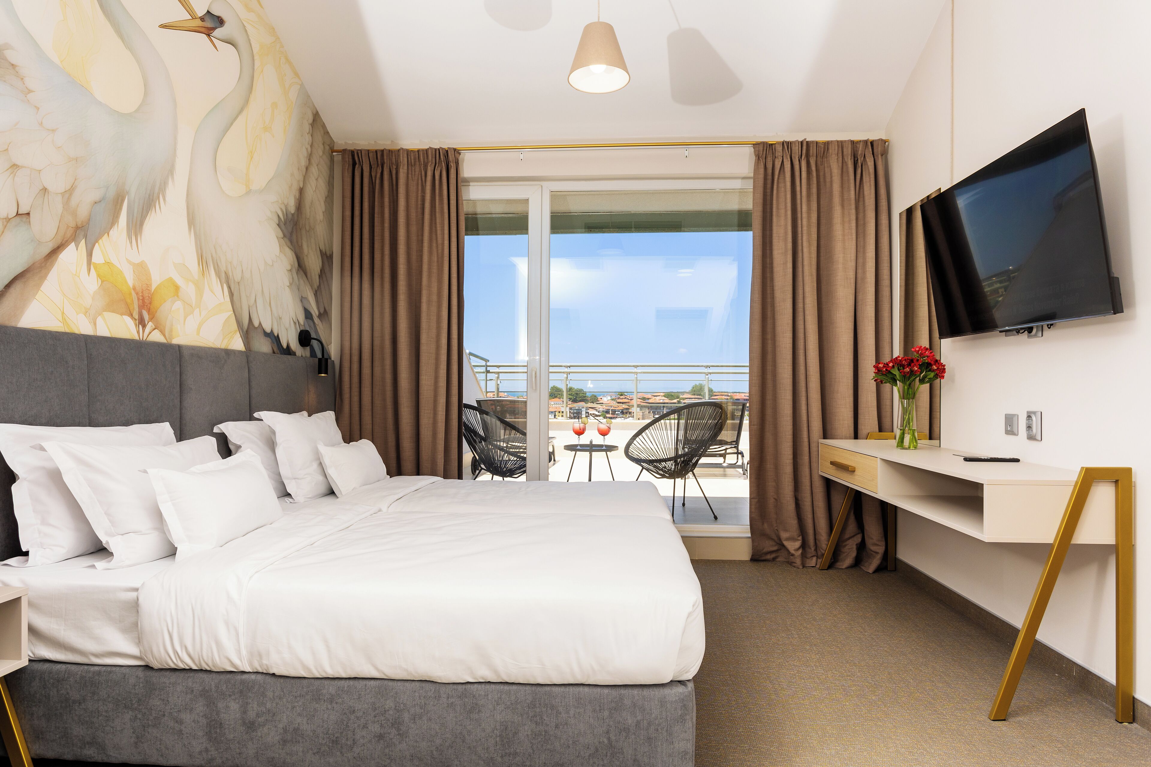A hotel room with a bed, a desk, a TV, and a balcony with chairs.