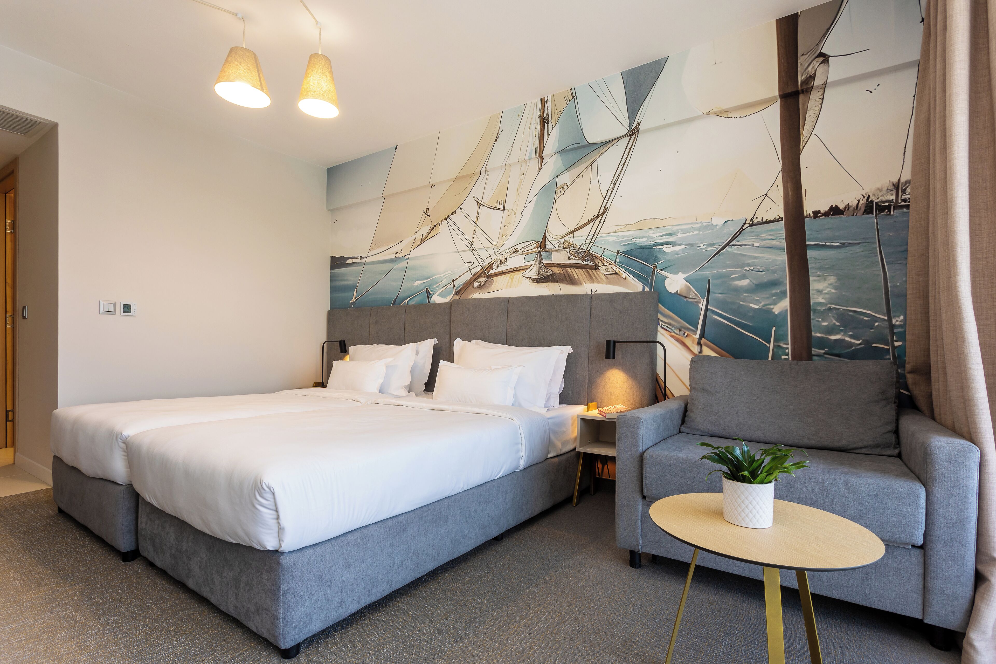A modern hotel room with a large bed, a grey sofa, a small round table, and a wall mural of sailboats.